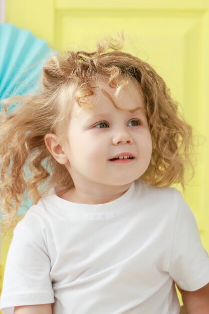 Retrato de niña rizada rubia bastante pequeña en camiseta blanca sonriendo sobre fondo amarillo.