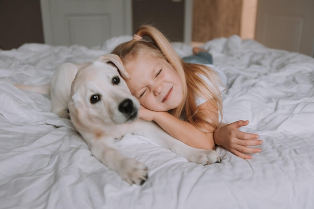 Retrato de una niña rubia acostada en la cama con su amado perro en los brazos. una chica con una camiseta blanca y jeans y una mascota yacen sobre ropa de cama blanca. espacio para texto. foto de alta calidad