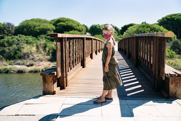 Retrato de niña rubia de ojos azules con mascarilla de vacaciones en un puente de madera con pinos con rostro serio con vestido verde en medio de la pandemia de coronavirus