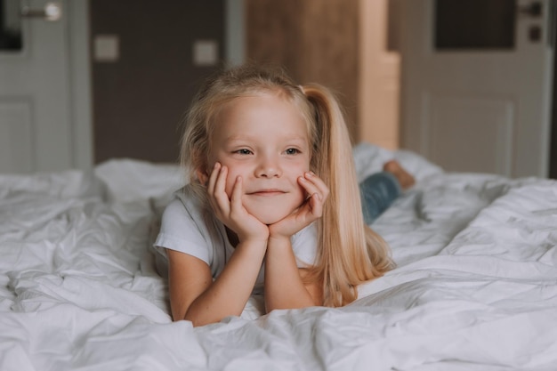 Retrato de una niña rubia sonriente con una camiseta blanca y jeans acostados boca abajo en la cama con ropa de cama blanca. recreación infantil. espacio para texto. foto de alta calidad