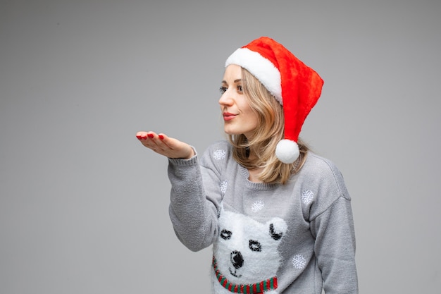 Retrato de niña rubia en suéter gris y rojo gorro de Papá Noel enviando aire beso con la mano