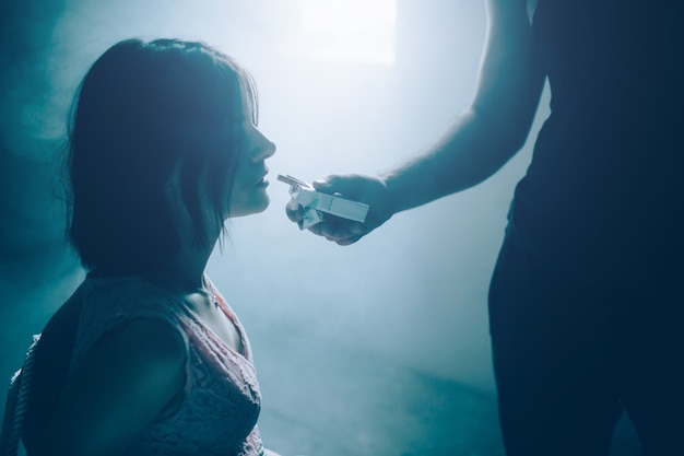 Retrato de niña sentada atada y mirando una pequeña caja de cigarrillos que el tipo de camisa negra está sosteniendo frente a ella