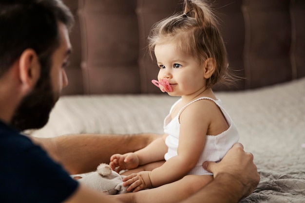 Retrato de una niña sentada en una cama en un dormitorio y mirando a su padre