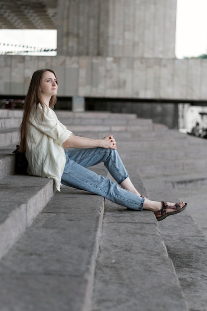 Retrato de niña sentada en las escaleras. Mujer moderna en jeans y camisa descansando en la calle de la ciudad.