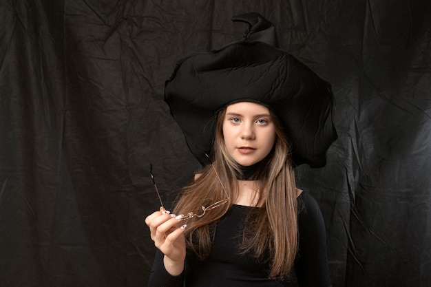 Foto retrato de niña en sombrero de bruja puntiagudo con gafas en sus manos sobre fondo negro disfraz de halloween