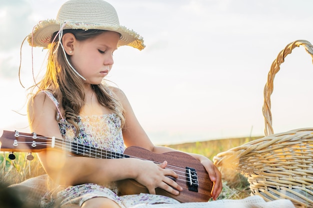 Retrato de una niña con sombrero sentada en una manta en un campo de heno seco con un picnic aprendiendo a tocar el ukelele Vista lateral