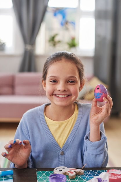 Retrato de niña sonriendo a la cámara sentado en la mesa y sosteniendo el huevo de Pascua que se prepara para la Pascua en casa