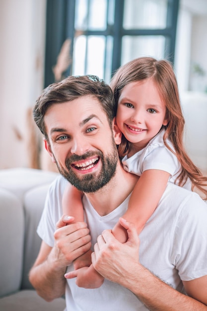 Retrato de una niña sonriente abrazando a un padre feliz en casa mirando la cámara