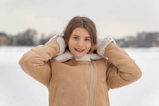 Retrato de niña sonriente en abrigo de piel de oveja beige corrigiendo el fondo del terreno nevado del cabelloMujer feliz en ropa de invierno