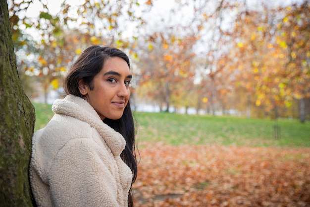 Retrato de una niña sonriente apoyada con la espalda en un árbol en un parque público durante un día de otoño con hojas de naranja caídas en el fondo