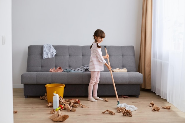 Retrato de una niña sonriente y feliz lavando el suelo con un trapeador en casa limpiando la sala de estar de pie en un lío ayudando a la madre con las tareas domésticas