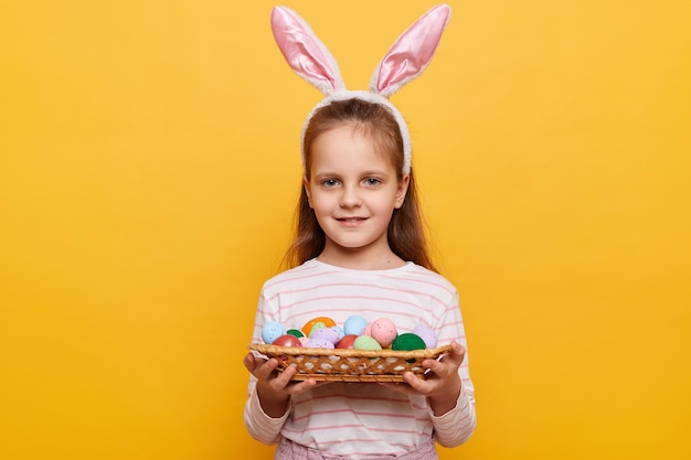 Retrato de una niña sonriente con orejas de conejo en la cabeza con una cesta de huevos de colores en las manos posando aislada en un fondo amarillo celebrando las vacaciones