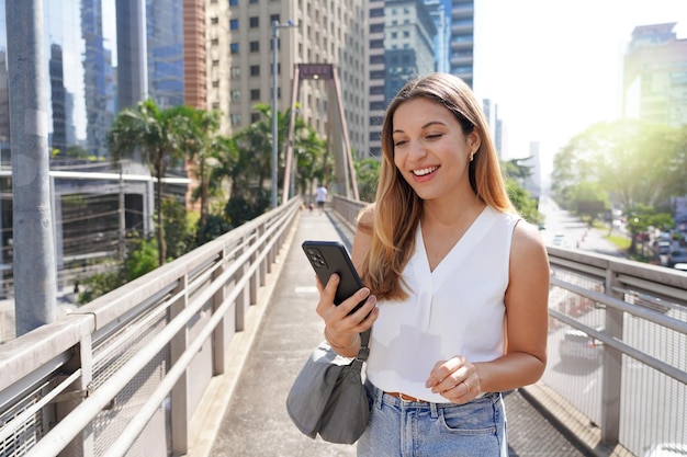 Retrato de niña sonriente en la pasarela viendo su smartphone en la moderna metrópolis sostenible de Sao Paulo, Brasil