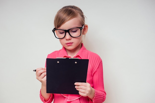 Retrato de niña sonriente pensativa en una blusa rosa y gafas sosteniendo cuaderno y lápiz con tableta copyspace. Niño genio con la idea sobre fondo blanco en el espacio de copia de estudio