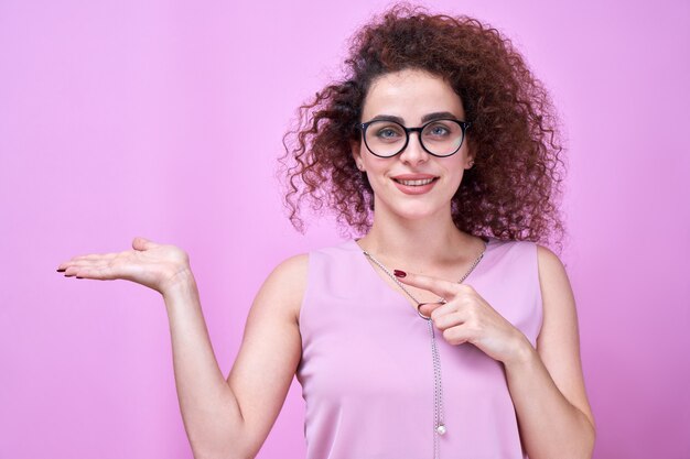 Retrato de niña sonriente rizada caucásica pretendiendo sostener algo en la palma y apuntando con un dedo aislado sobre fondo de estudio rosa con espacio de copia