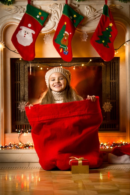 Retrato de niña sonriente sentada en una bolsa roja para regalos en la chimenea