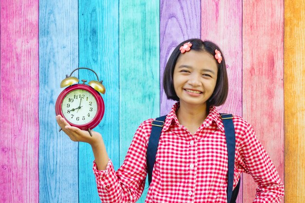 Foto retrato de una niña sonriente sosteniendo un viejo despertador mientras está de pie contra una pared de varios colores