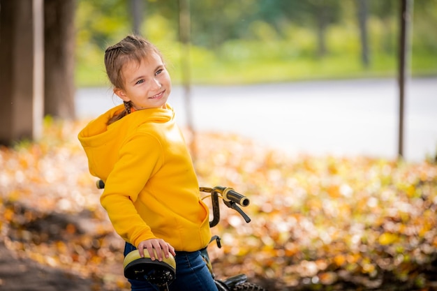 Retrato de una niña sonriente en una sudadera amarilla con bicicleta en un parque de otoño agitando la mano amistosa
