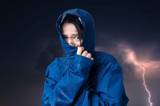 Retrato de una niña sonriente vestida con impermeable azul en gotas posando con capucha contra el fondo de un cielo sombrío con relámpagos.