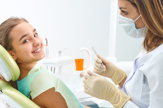 Retrato de una niña con sonrisa toothy sentado en el sillón dental con el médico en el fondo en el consultorio dental