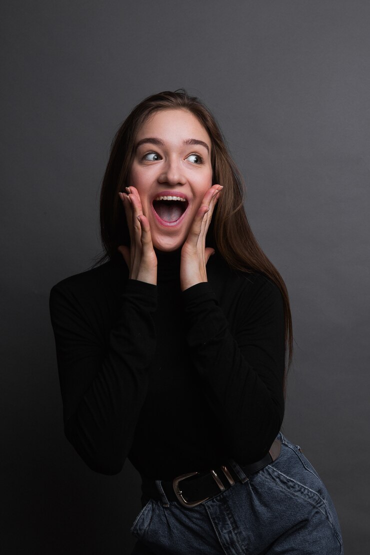 Retrato de niña sorprendida en camisa negra sobre fondo gris oscuro. Mujer joven sorprendida