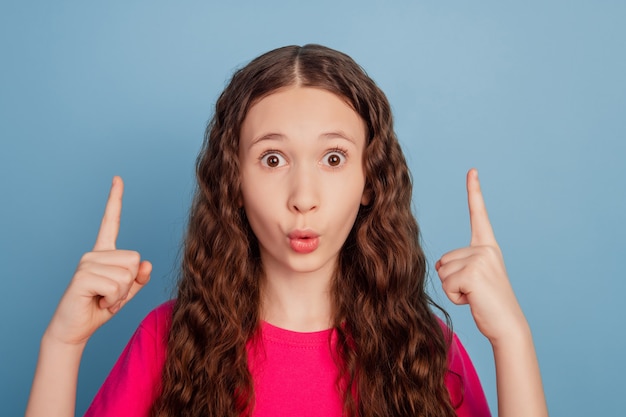 Retrato de una niña sorprendida y divertida con los dedos directos del espacio vacío con la boca abierta sobre fondo azul.