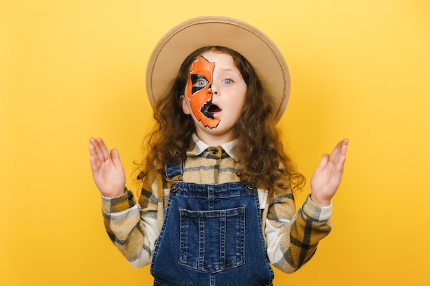 Foto retrato de niña sorprendida con máscara de maquillaje de halloween lleva sombrero mirando a la cámara con asombro, abriendo los ojos y la boca con sorpresa, posando aislado sobre fondo de color amarillo en estudio