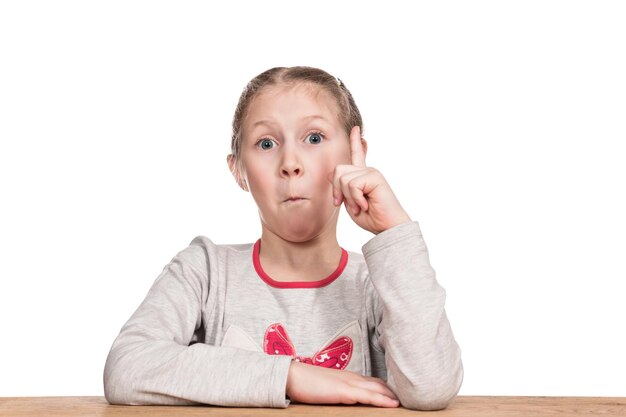 Retrato de una niña sorprendida sentada en una mesa con un dedo levantado aislado en un fondo blanco