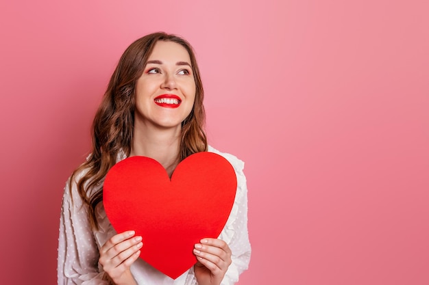 Retrato de una niña sosteniendo una gran tarjeta de corazón rojo aislada en un fondo rosa