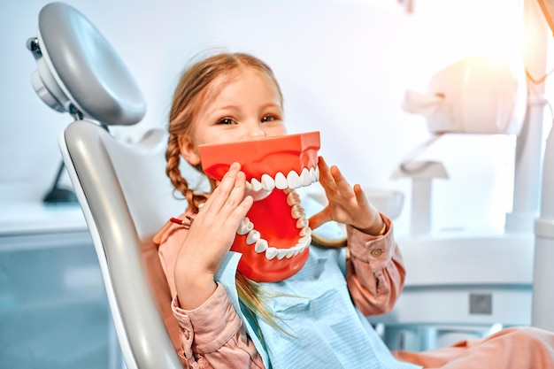 Retrato de una niña sosteniendo un modelo de mandíbula con dientes mientras está sentada en un consultorio dental mirando la cámara y sonriendo Odontología infantil Copiar espacio