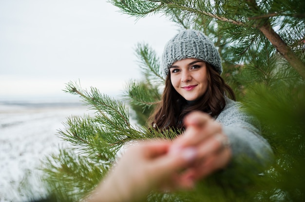 Retrato de niña suave en abrigo gris y sombrero contra el árbol de año nuevo al aire libre sosteniendo la mano.