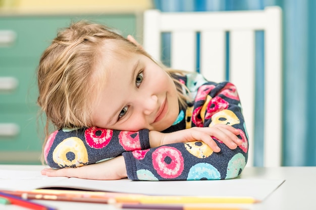 Retrato de niña en suéter colorido sentado en el escritorio y apoya la cabeza en las manos