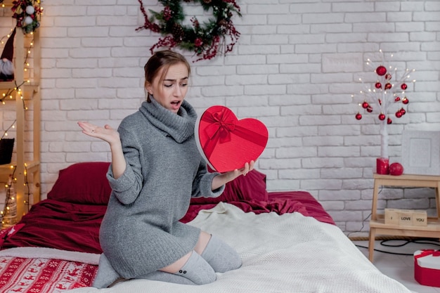 Retrato de una niña en un suéter gris con una caja de regalo roja en forma de corazón sentado en el apartamento, concepto de San Valentín, espacio de copia