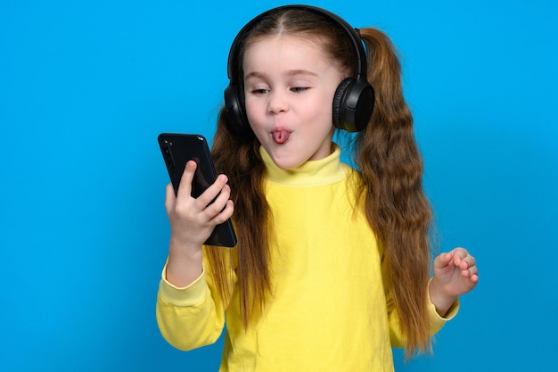 Retrato de una niña con un teléfono y auriculares inalámbricos un niño en un suéter amarillo sobre un fondo azul.