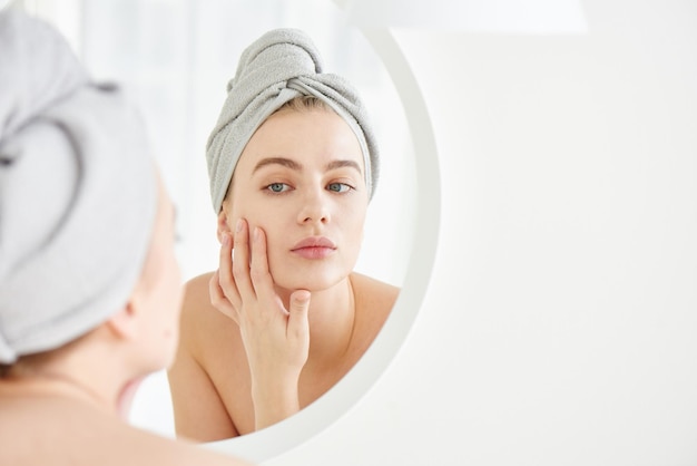 Retrato de niña con toalla en la cabeza en el baño blanco mira y toca su rostro en el espejo y disfruta de la juventud y la hidratación Belleza natural cuidado en el hogar para la piel problemática