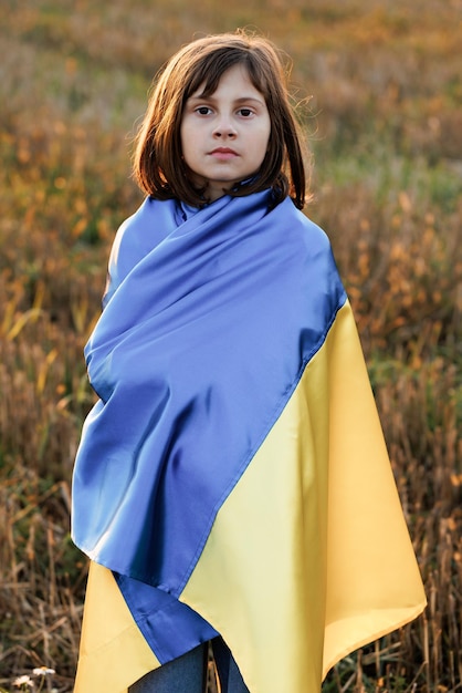 Retrato de niña ucraniana triste se encuentra con una bandera de ucrania en medio del campo contra una puesta de sol