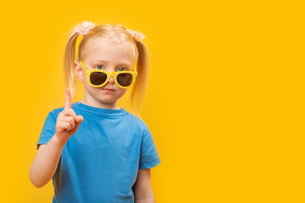 Retrato de niña viste camiseta azul y gafas de sol amarillas sobre fondo amarillo El niño levantó su dedo índice Maqueta de espacio de copia