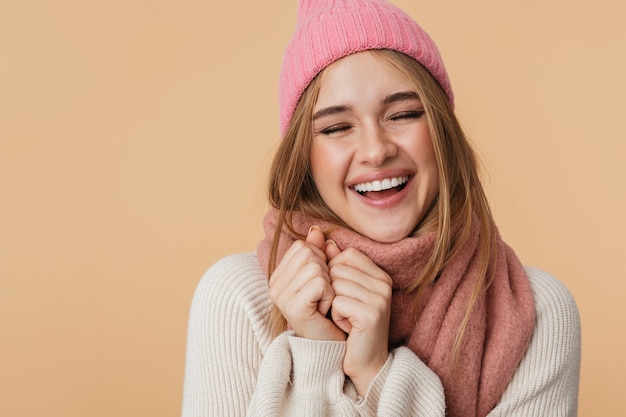 Retrato de niña vistiendo gorro y bufanda de invierno sonriendo y presionando sus manos en el pecho aislado en beige