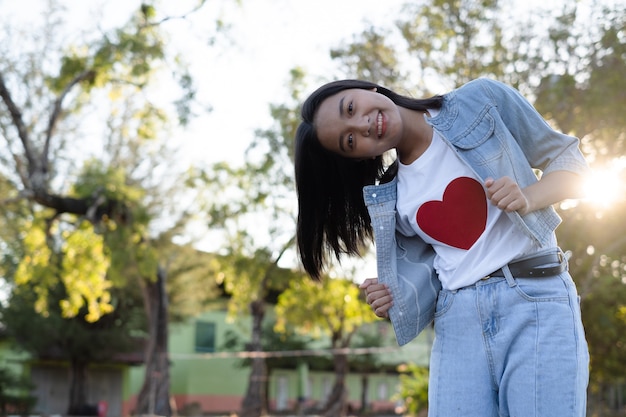 Retrato de niña vistiendo jean y camisa blanca con corazón rojo sobre fondo de naturaleza.