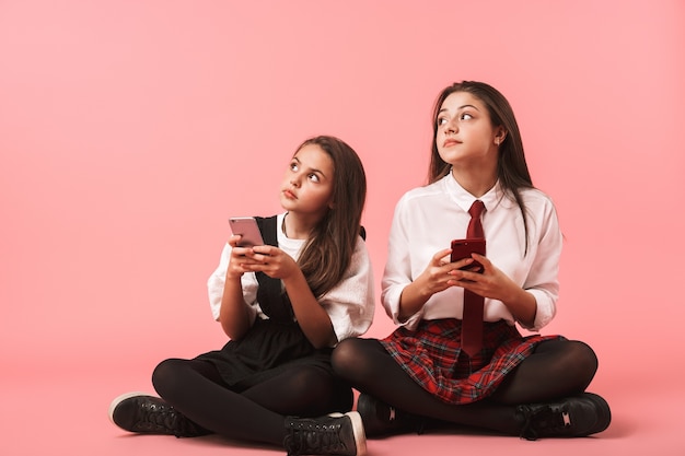 Retrato de niñas alegres en uniforme escolar usando teléfonos móviles, mientras está sentado en el piso aislado sobre la pared roja