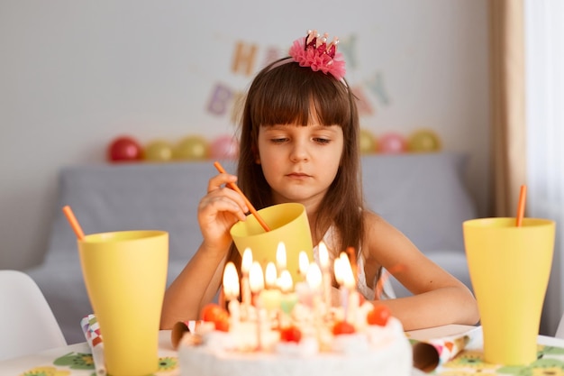 Retrato de una niñita aburrida de cabello oscuro con flores en el pelo sentada en la mesa con pastel de cumpleaños posando con un vaso de limonada se ve triste y molesta