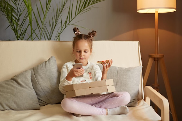 Retrato de una niñita graciosa y hambrienta con ropa informal sentada en un sofá comiendo pizza y sosteniendo un teléfono móvil tomando un refrigerio viendo dibujos animados mientras revisa las redes sociales