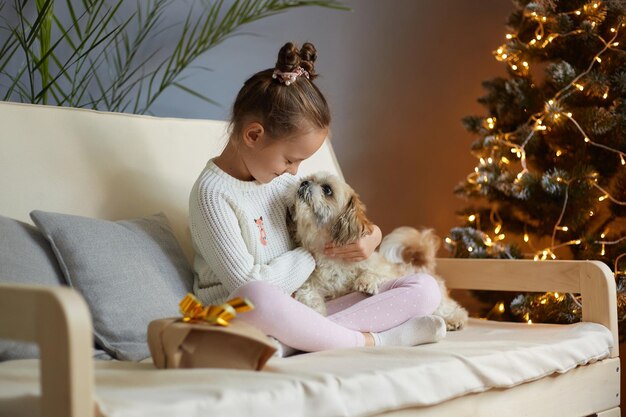 Retrato de una niñita de pelo oscuro con dos bollos de pelo con ropa informal sentada en un sofá con su perro mirando a un cachorro con amor y celebrando el año nuevo con su adorable mascota