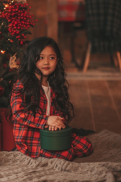 Retrato de una niñita sonriente asiática sincera en pijama de cuadros rojos sentada con regalos en casa de Navidad