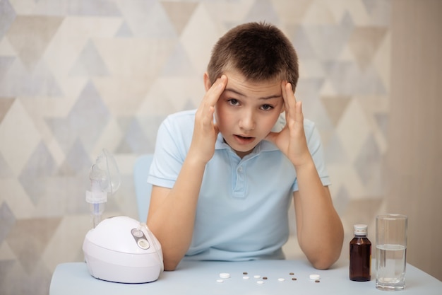 Retrato de un niño, un adolescente sentado en una mesa con pastillas y un inhalador, tratamiento en el hogar