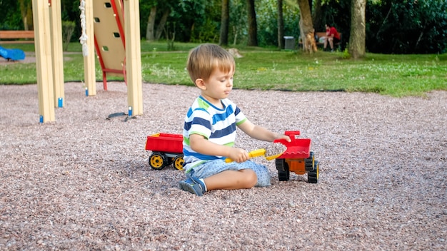 Retrato de niño adorable niño de 3 años jugando con camión de juguete con remolque en el patio de recreo en el parque. Niño cavando y construyendo con arena.