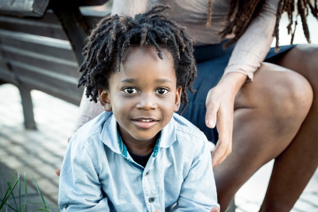 Retrato de niño africano con madre sonriendo en el parque. Niño en peinado de deadlocks jugando en el parque con su madre. Retrato de niño negro sonriente con madre sentada en un banco en el jardín