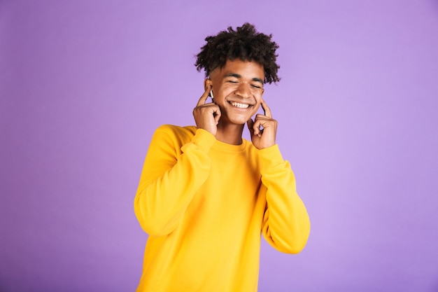 Retrato de niño afroamericano satisfecho con elegante peinado afro sonriendo con los ojos cerrados, mientras escucha música a través del auricular bluetooth, aislado sobre fondo violeta