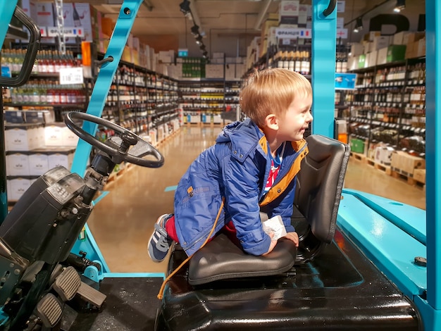 Retrato de niño alegre sentado en el asiento del conductor de la carretilla elevadora en el almacén o tienda grande con estantes altos con alimentos y bebidas