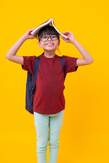 El retrato del niño asiático divertido de la muchacha con el libro en la cabeza que sonríe, estudiante tailandés bonito en camisa roja con los vidrios tiene posición y mirada atractivas, conocimiento y sabiduría